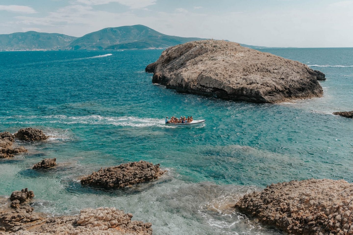 Small white boat of people driving through turquoise water with islands in the distance. Bisevo Island, Croatia.