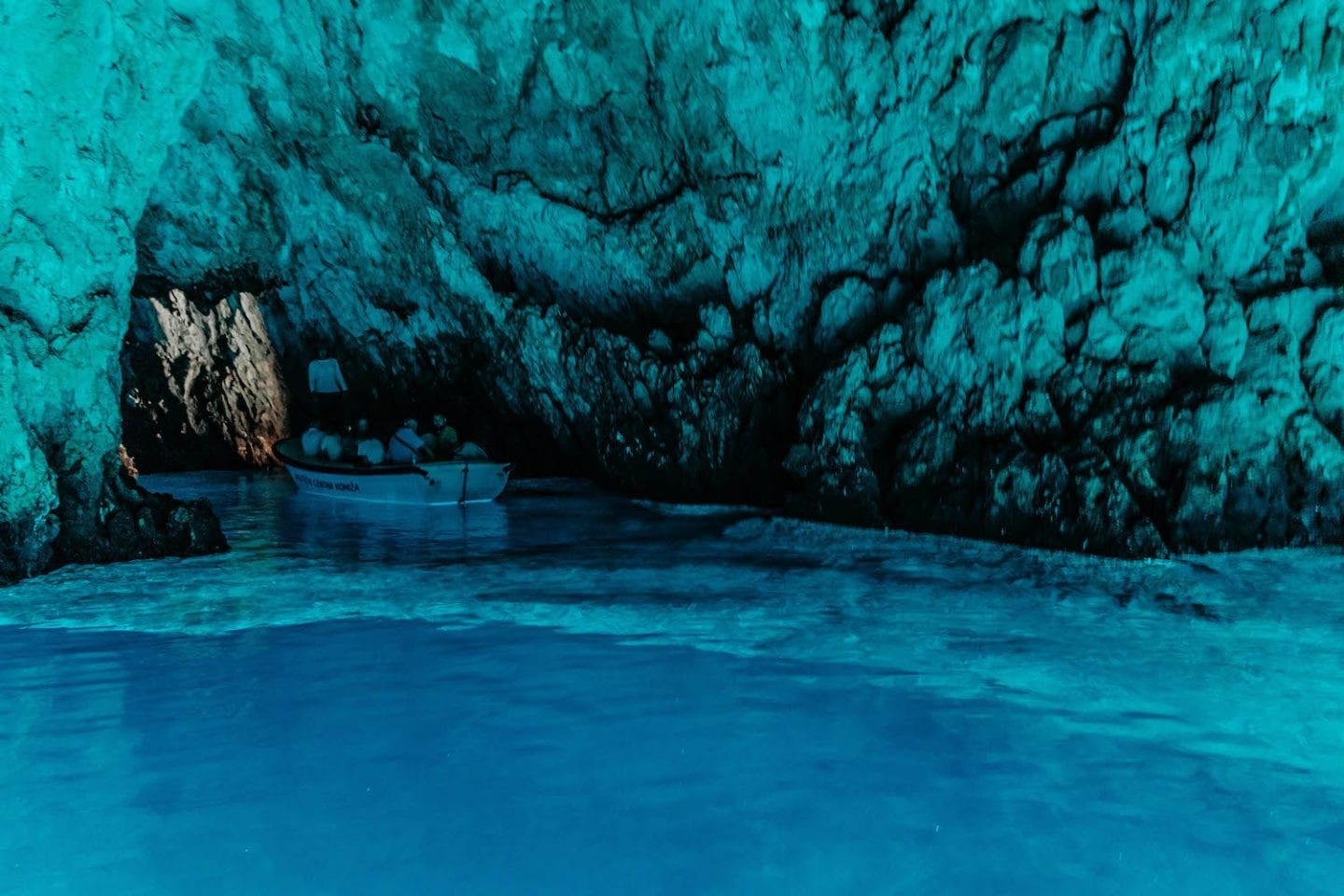 Dark cave lit up by blue water with small wooden boat of people drifting through the cavern - Blue Cave in Croatia