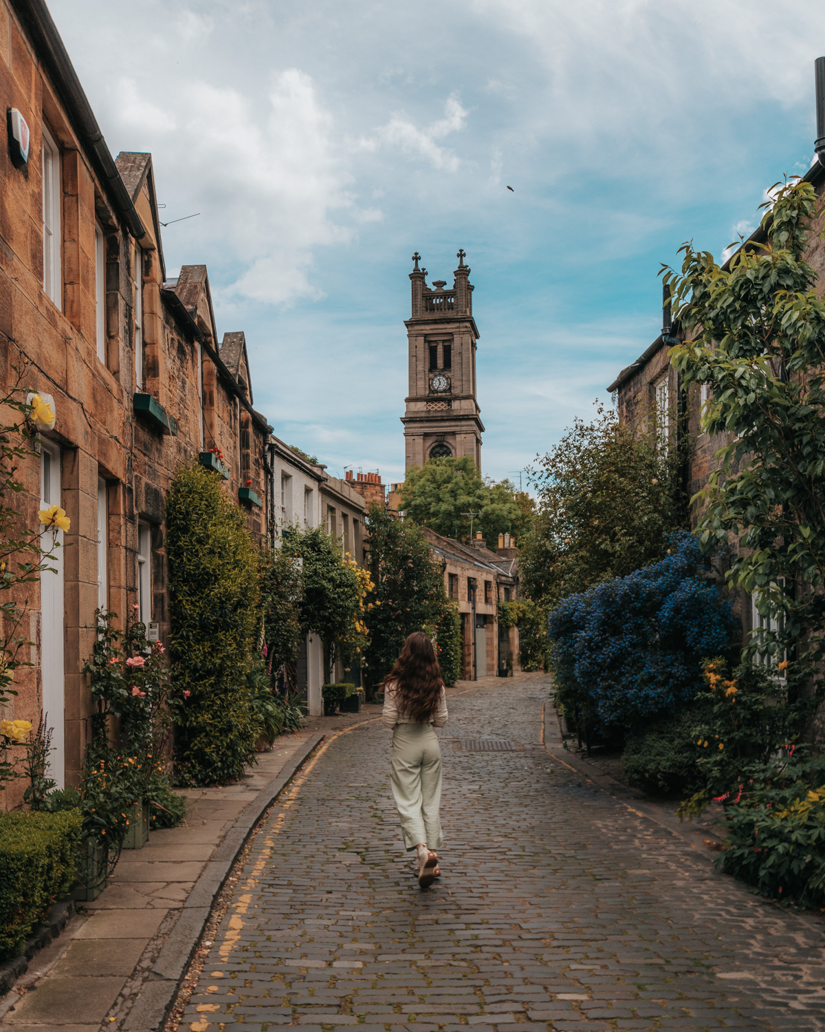 Girl standing in the middle of a cobblestone street lined with small houses and flowers with a church tower in the background - Circus Lane, Edinburgh, Scotland