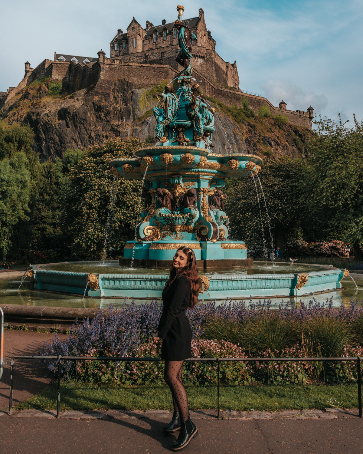 Girl in black dress standing in front of blue and gold fountain below a castle - Ross Fountain in Edinburgh, Scotland
