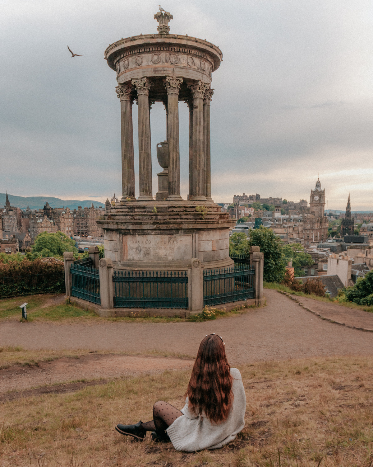 Girl sitting on slight hill in front of a monument with city skyline in background - Calton Hill, Edinburgh, Scotland