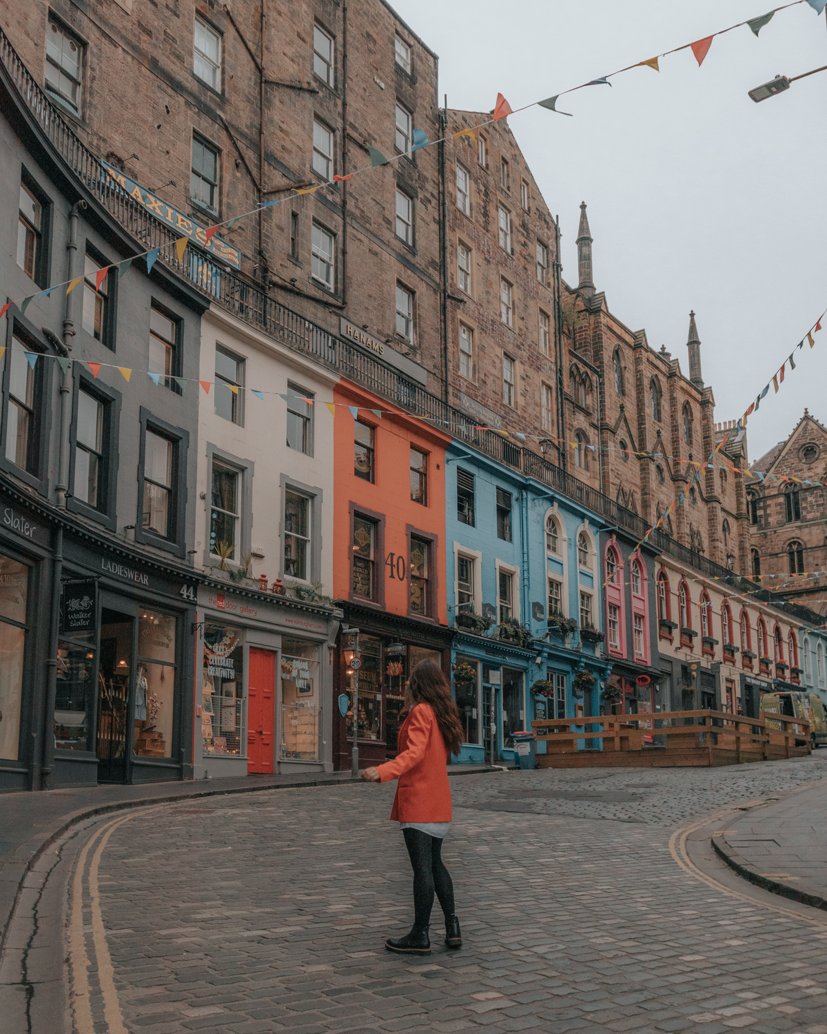 Girl in red jacket standing in front of a winding street with colorful storefronts - Victoria Street in Edinburgh, Scotland