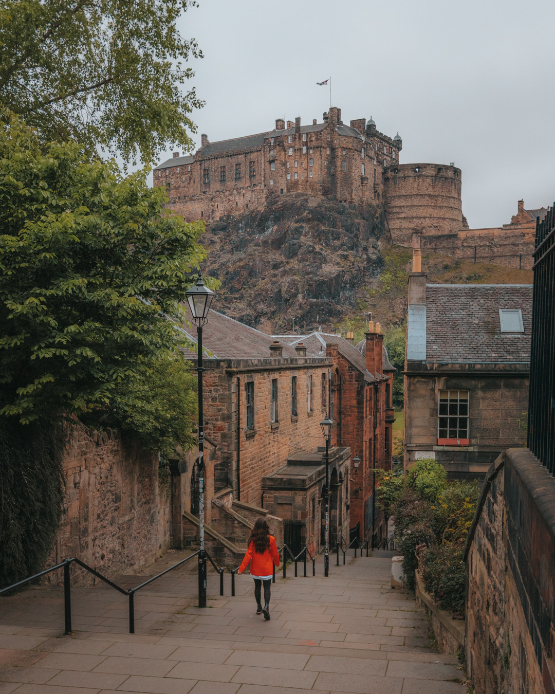 Girl in bright red jacket standing at the top of steps with a view of an ancient castle on a hill in the background - The Vennel in Edinburgh, Scotland