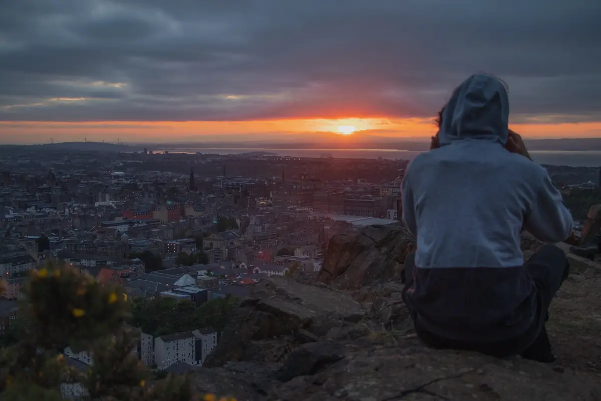 Man sitting on cliff edge during sunset overlooking historic city - Salisbury Crags in Edinburgh, Scotland